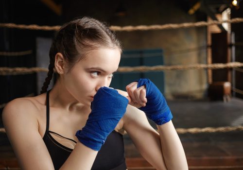 Martial arts, boxing, kickboxing and training concept. Close up portrait of beautiful teenage girl exercising indoors, wearing handwraps, holding clenched fists at her face, preparing to deliver punch